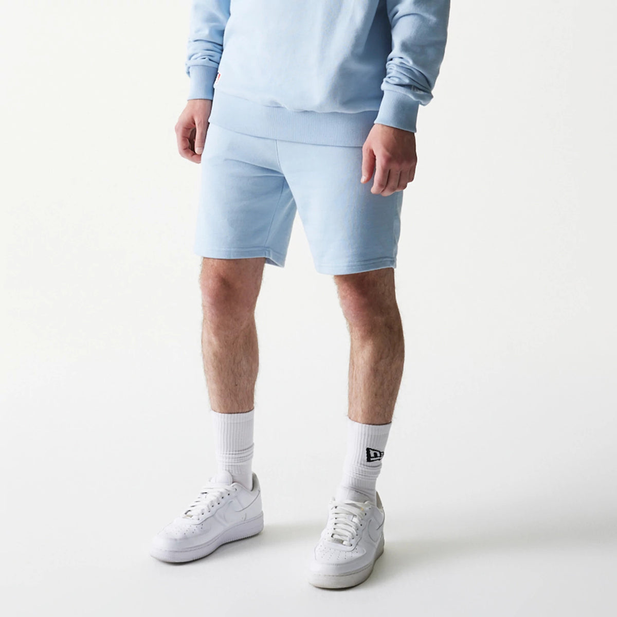 New Era New York Yankees MLB League Essential Midi Blue Shorts, front view on-model showing the light blue shorts with a straight hem above the knee, paired with white trainers and socks featuring a printed New Era logo.