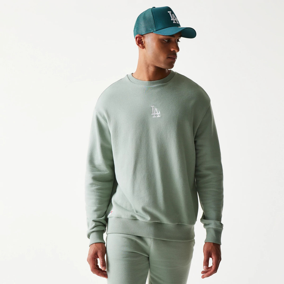 New Era LA Dodgers MLB Midi Green Oversized Crew Neck Sweater, front view on a model with a matching green cap, featuring a white embroidered LA logo on the chest and ribbed cuffs and hem.