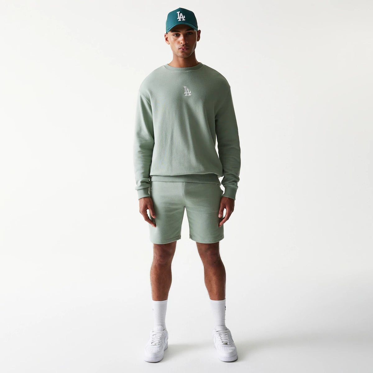 New Era LA Dodgers MLB League Essential Midi Green Shorts, front view on-model showing the knee-length green shorts paired with a matching sweatshirt, LA logo on the left side of the shorts, styled with white trainers and socks.