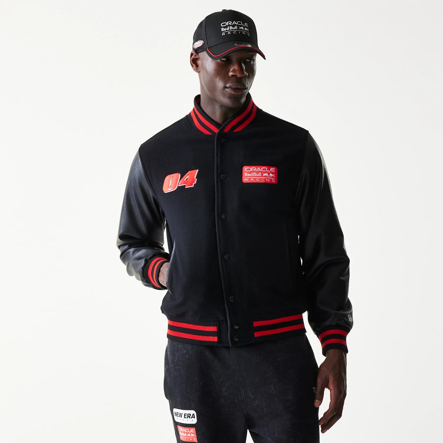 New Era Red Bull Racing Black Varsity Jacket, front view on-model showing an embroidered number 04 on the left chest, a Red Bull Racing patch on the right chest, and red and black striped cuffs and hem with a snap button closure.