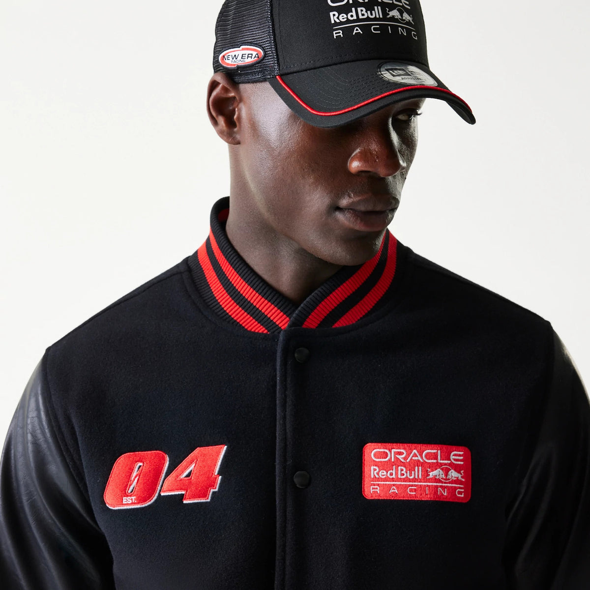 New Era Red Bull Racing Black Varsity Jacket, front view on a model showing red embroidered 04 and Oracle Red Bull Racing patches with a red striped collar, paired with a matching cap displaying the New Era logo on the side.