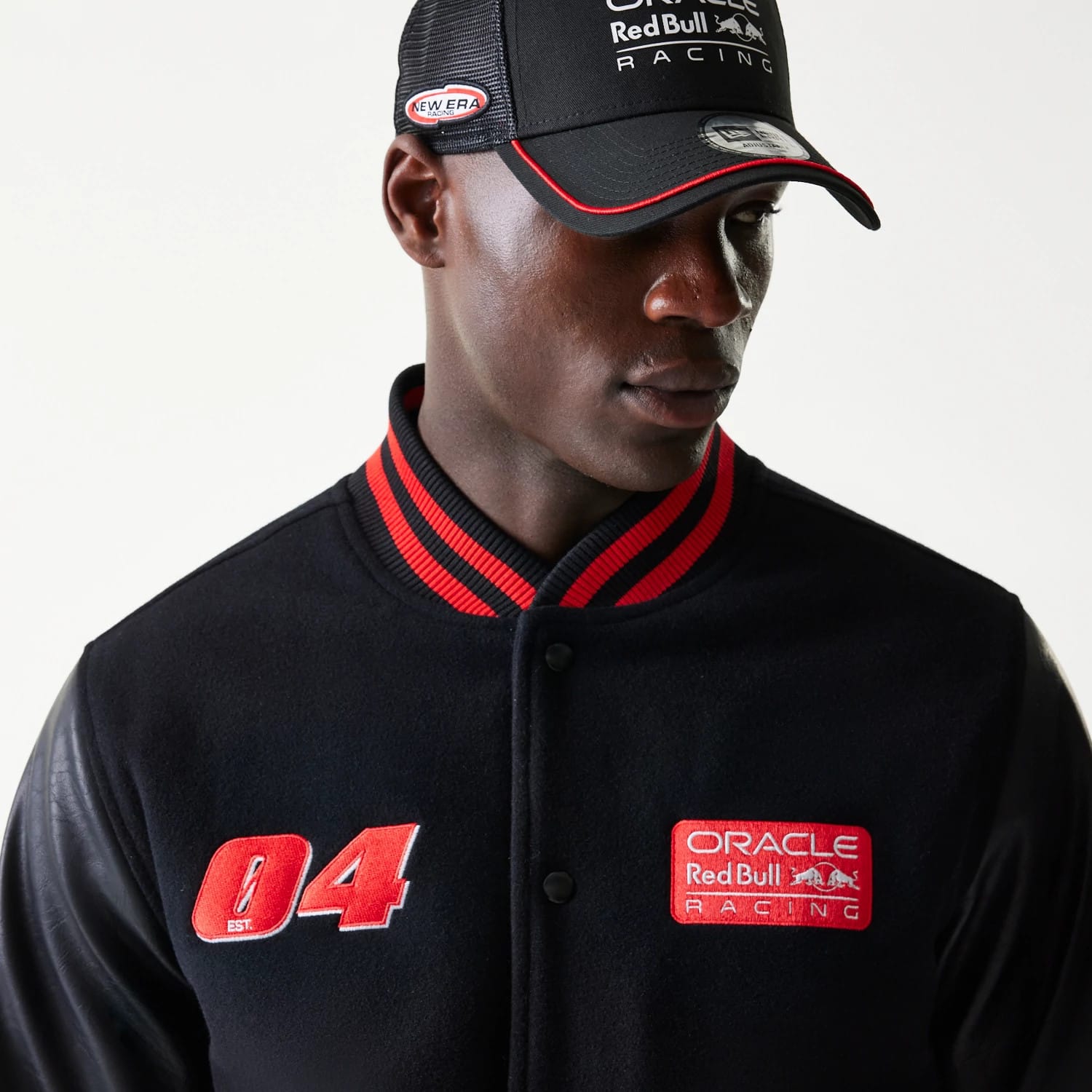 New Era Red Bull Racing Black Varsity Jacket, front view on a model showing red embroidered 04 and Oracle Red Bull Racing patches with a red striped collar, paired with a matching cap displaying the New Era logo on the side.