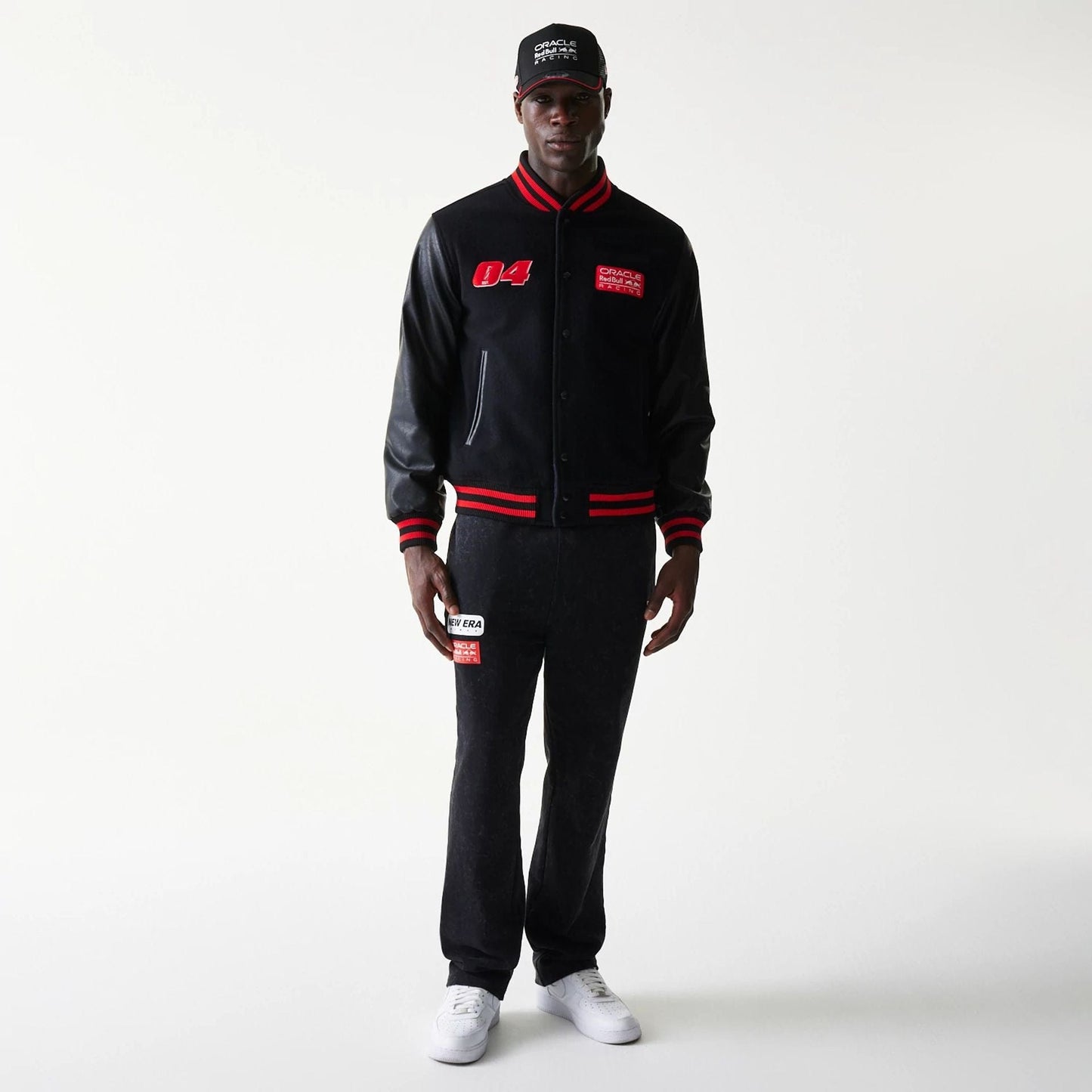 New Era Red Bull Racing Black Varsity Jacket, front view on-model displaying red embroidered 04 on the right chest, a red patch on the left chest, striped red and black cuffs and hem, matched with a black cap and trousers.