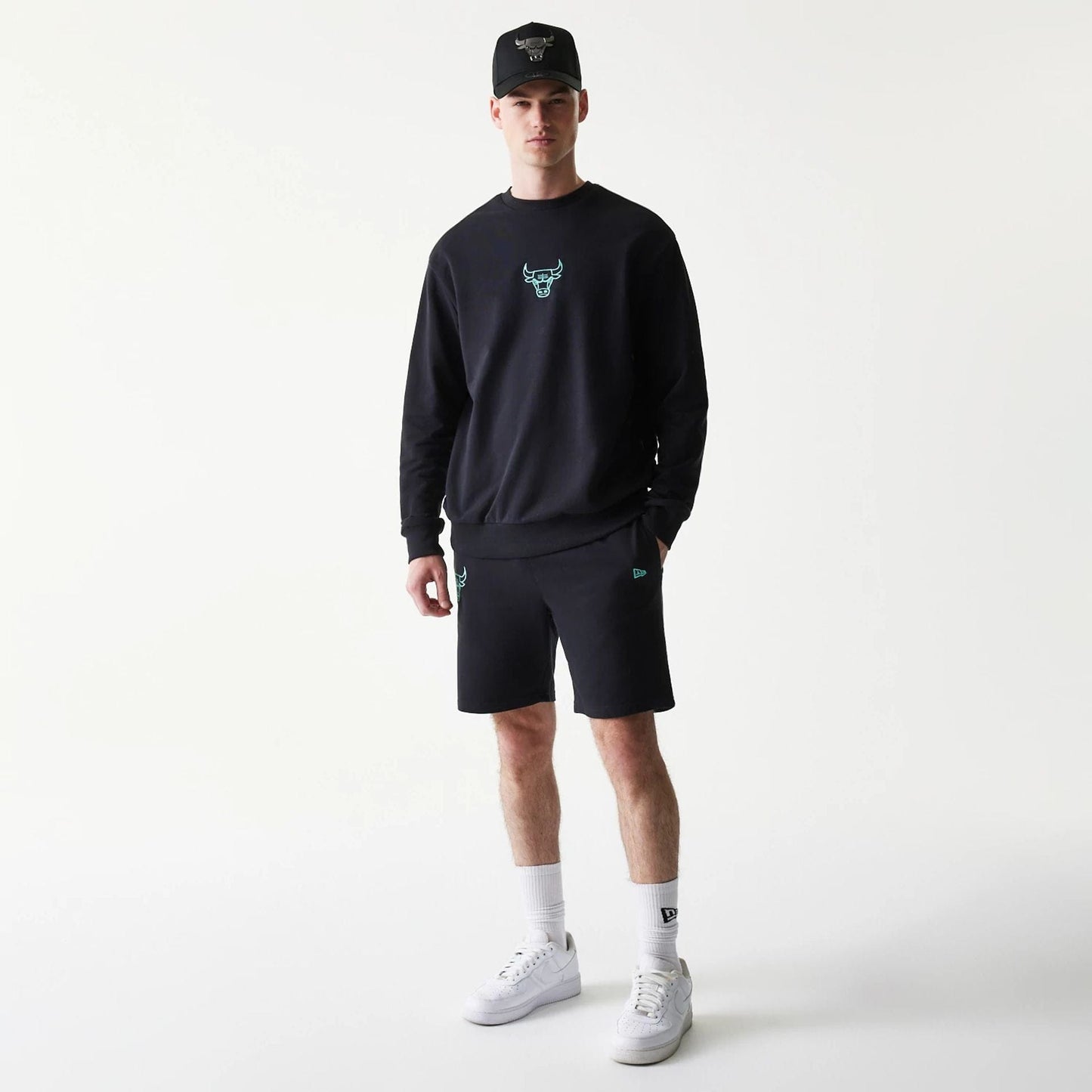 New Era Chicago Bulls NBA Core Black Shorts, front view on-model showing an embroidered teal bulls logo on the left leg, complemented by a matching sweatshirt, New Era logo on the right leg, and a cap styled for a cohesive look.