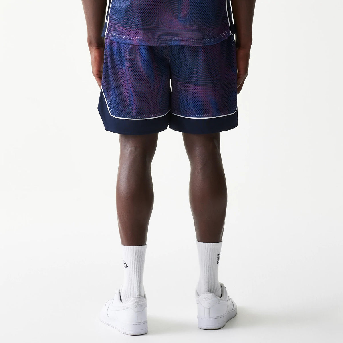 New Era Red Bull Racing Dark Blue Oversized Mesh Shorts, back view on a model, showcasing the dark blue mesh fabric with purple accents and white piping at the hem, matched with white trainers and socks for a sporty look.