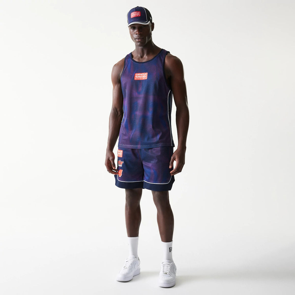 New Era Red Bull Racing Dark Blue Oversized Mesh Shorts, front view on a model wearing matching dark blue mesh tank, showing an orange-red printed logo on the left leg, with contrasting white trim outlining the hems and sides.