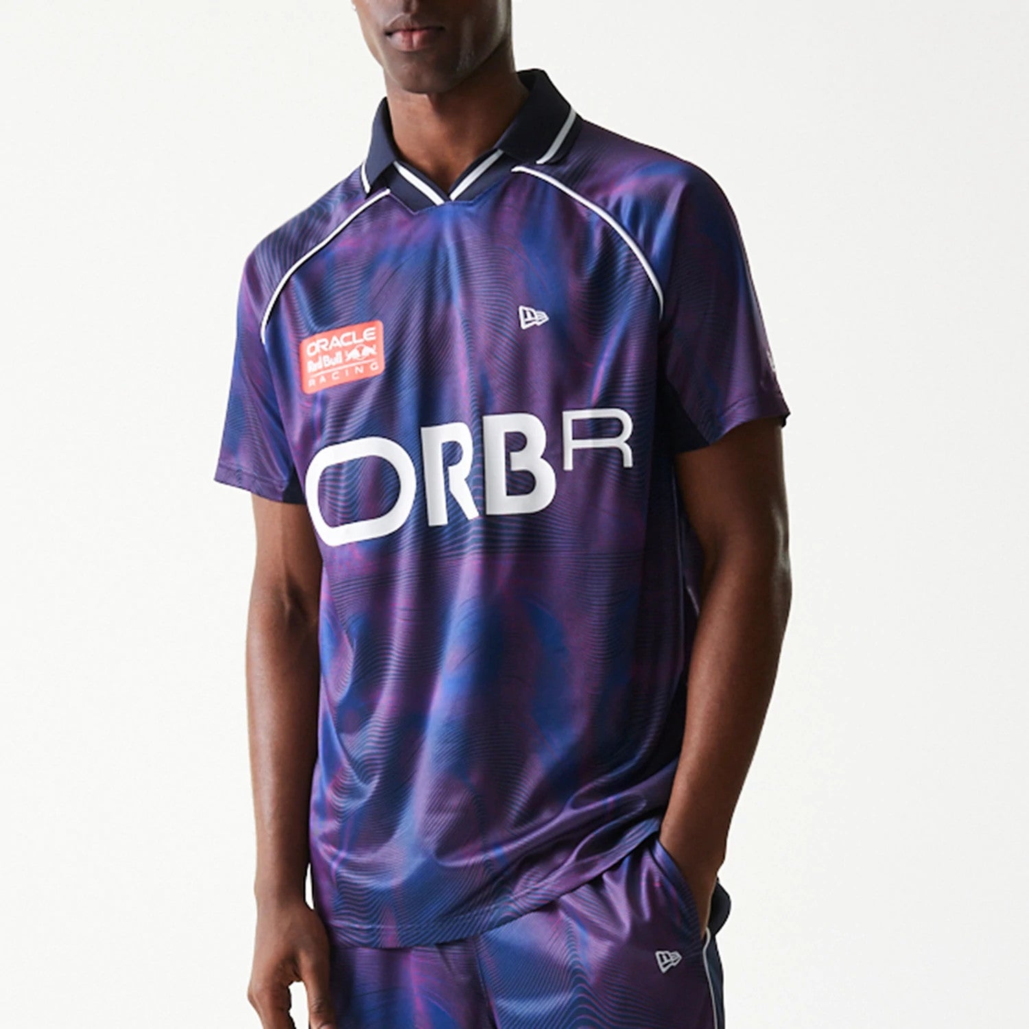 New Era Red Bull Racing All Over Print Dark Blue Football T-Shirt, front view on-model featuring a distinct all-over dark blue pattern with large white ORBR lettering, an embroidered New Era logo on the chest, and a contrast collar with white piping.