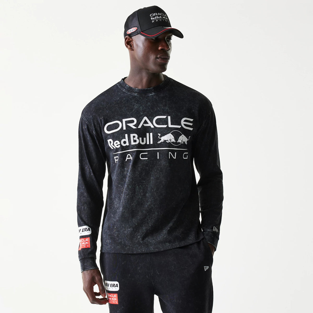 New Era Red Bull Racing Linear Graphic Black Long Sleeve T-Shirt, front view showing the white ORACLE Red Bull Racing logo across the chest, worn on-model with matching black trousers featuring logo patches and a cap with embroidered details.