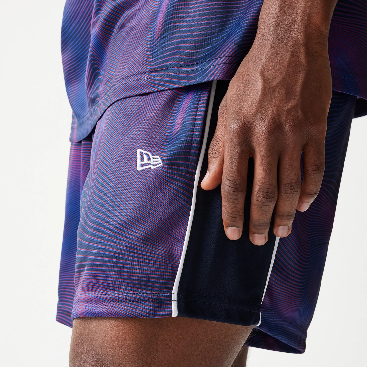 New Era Red Bull Racing All Over Print Dark Blue Football Shorts, side view showing the swirling blue and red pattern with white embroidered New Era logo near the hem, and vertical white piping along the edges.
