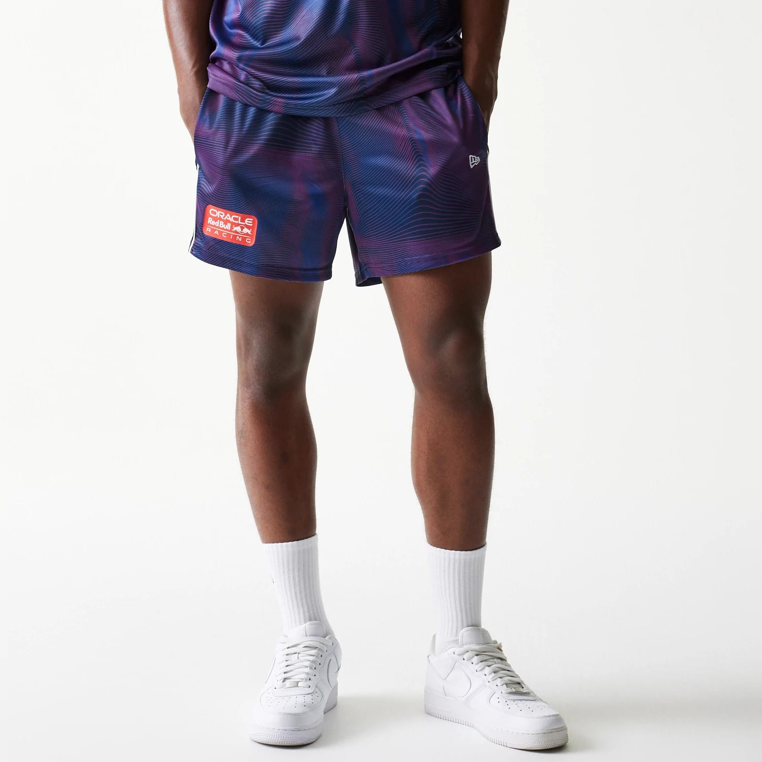 New Era Red Bull Racing All Over Print Dark Blue Football Shorts, front view on a model with white trainers showing a wave pattern, an embroidered New Era flag above the hem, and a red and white Red Bull Racing patch on the left leg.