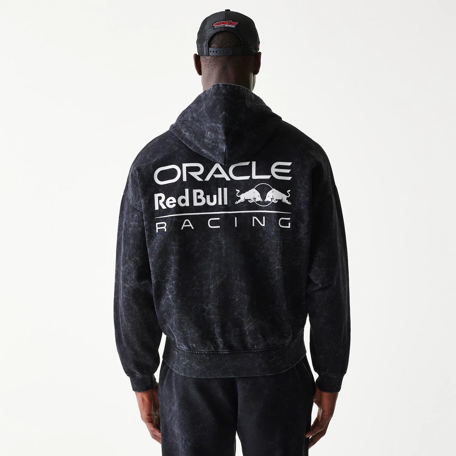 New Era Red Bull Racing Dropped Shoulder Black Dropped Shoulder Full Zip Hoodie, back view on a model styling matching trousers and cap, featuring large printed Oracle Red Bull Racing lettering across the upper back under the hood.