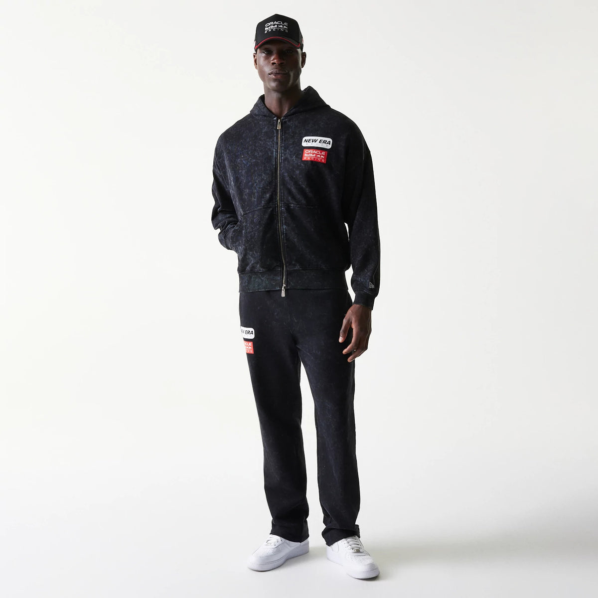 New Era Red Bull Racing Dropped Shoulder Black Dropped Shoulder Full Zip Hoodie, front view on a model styled with matching trousers and cap, featuring embroidered Red Bull and New Era patches on the chest and a full zip closure down the centre.