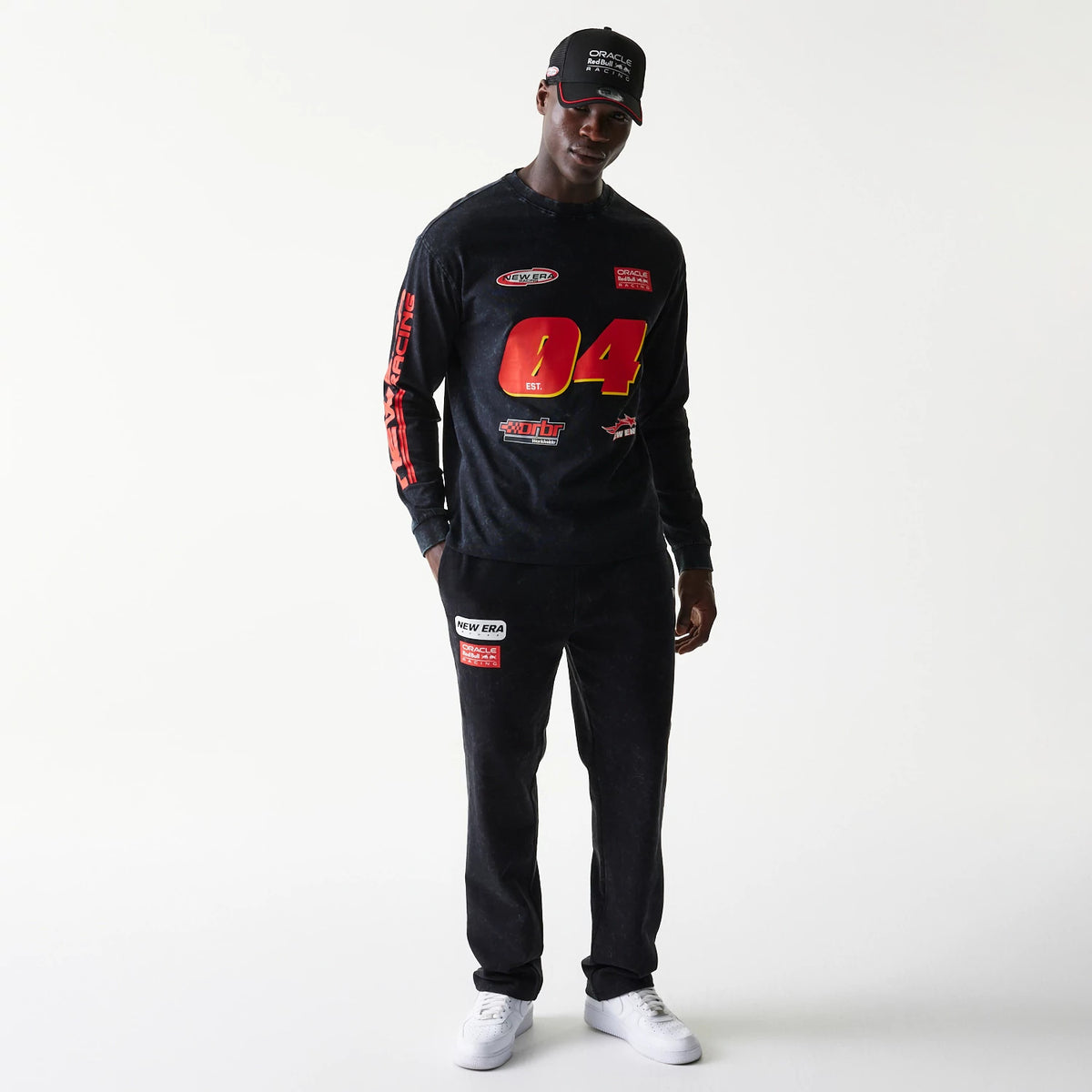 New Era Red Bull Racing Graphic Black Long Sleeve T-Shirt, front view on a model wearing matching loungewear, showcasing a red number 04, various racing logos, and Racing lettering down the sleeve, with white trainers completing the look.