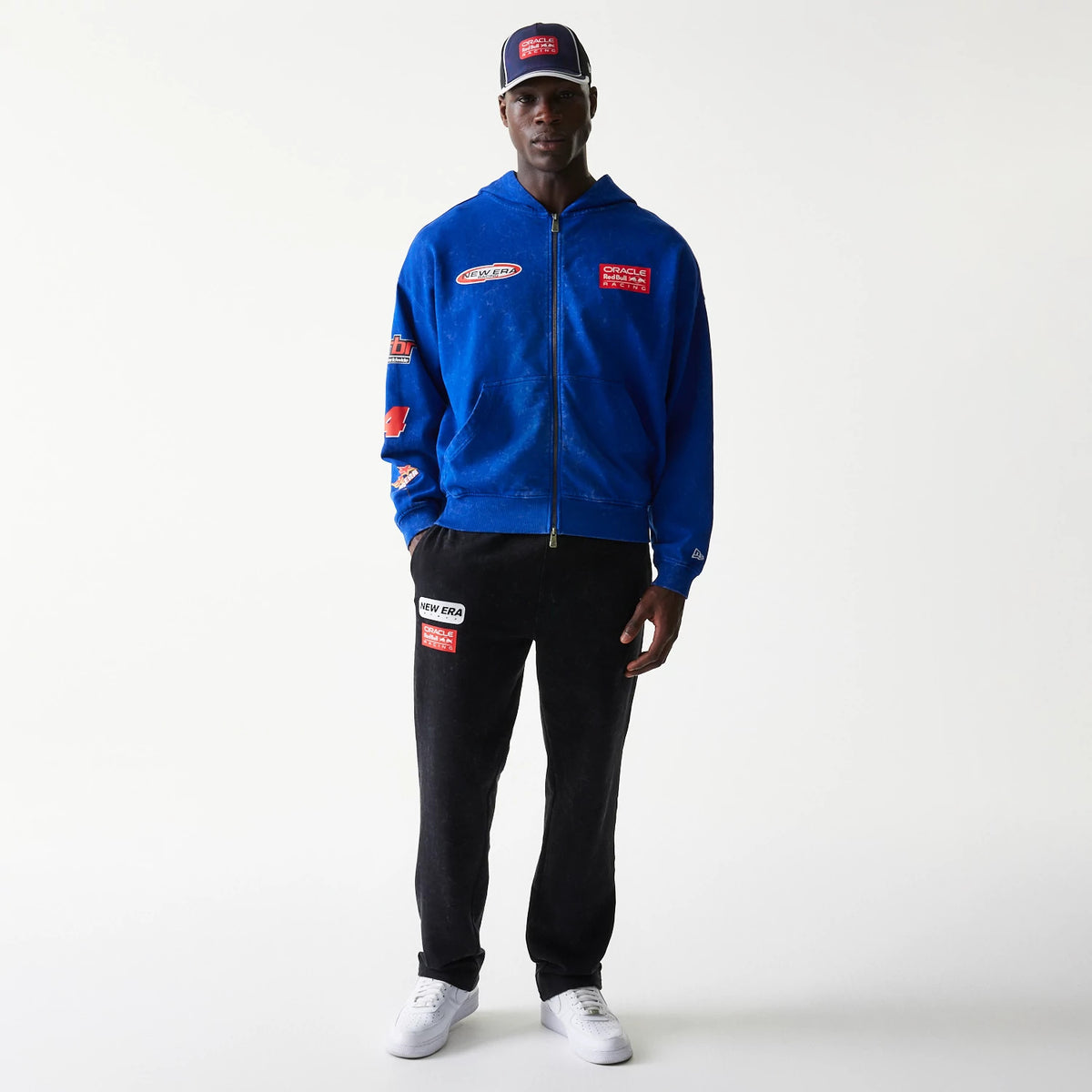 New Era Red Bull Racing Graphic Blue Dropped Shoulder Full Zip Hoodie, front view on a model with a matching cap, showing prominent team patches on the chest and sleeves, a zip closure, and a small embroidered New Era logo on the cuff.