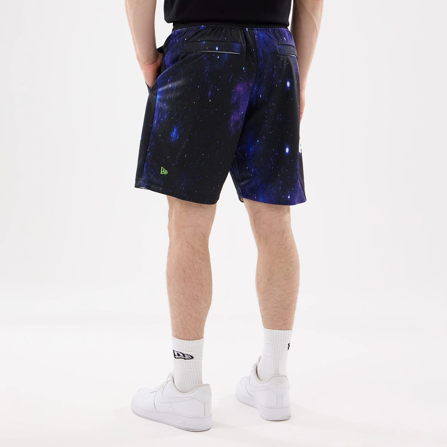 New Era Space Jam Warner Brothers Space Jam White Oversized Shorts, back view on-model showing a galaxy print design with black waistband and green embroidered New Era logo on the lower left, model styled with white trainers and socks.