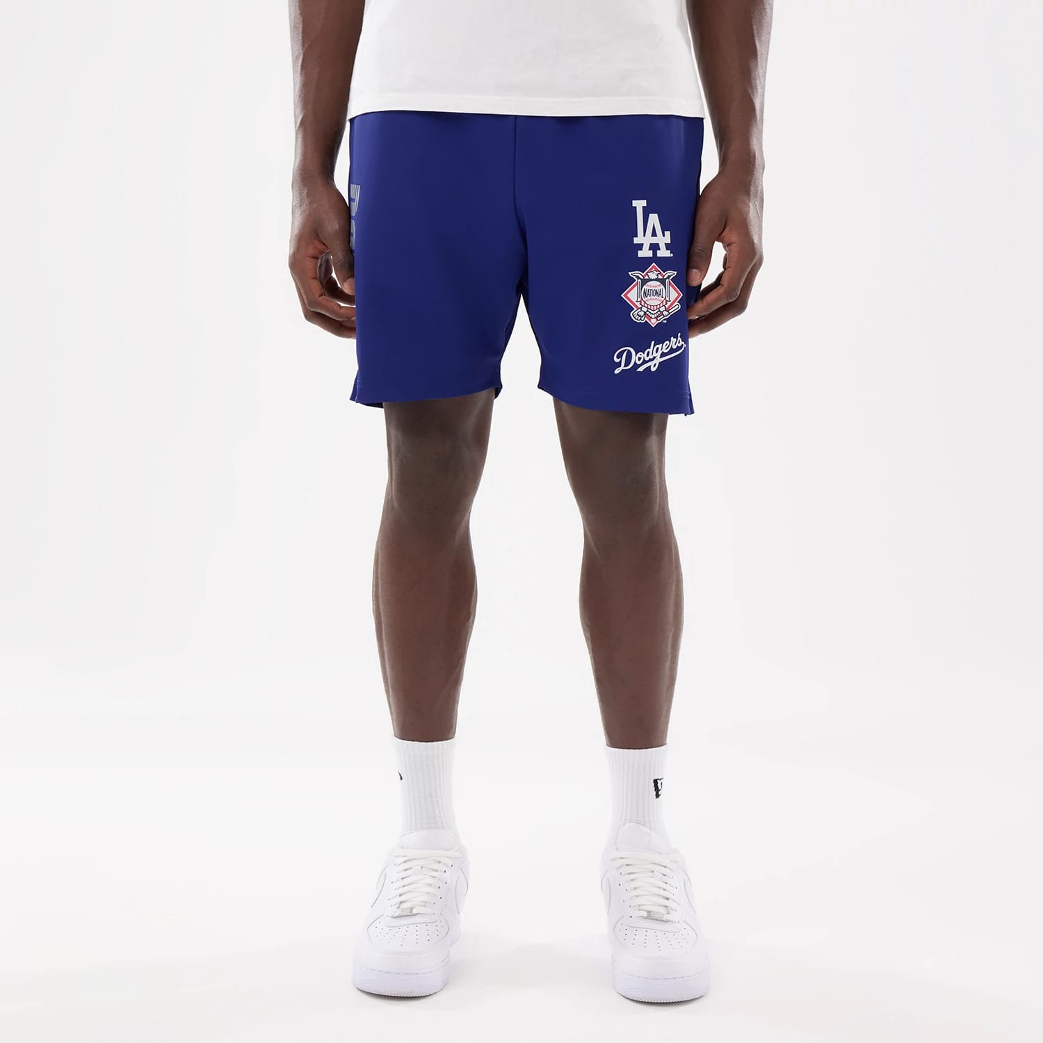 New Era LA Dodgers MLB Match Dark Blue Oversized Shorts, front view on-model showing white embroidered LA logo above a detailed MLB 75th-anniversary patch and Dodgers script, with the deep blue fabric standing out against white socks and shoes.