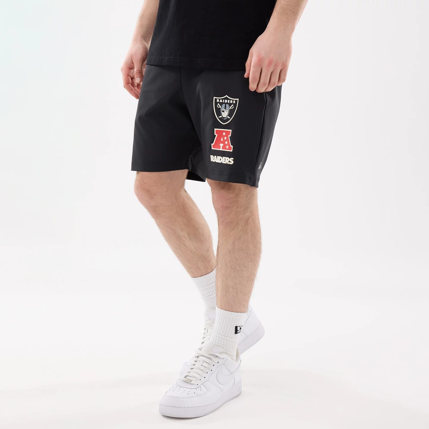 New Era Las Vegas Raiders NFL Match Black Oversized Shorts, front view on-model showing the black fabric with a printed Raiders shield logo and red AFC emblem on the right leg, white Raiders lettering below, styled with white trainers and socks.