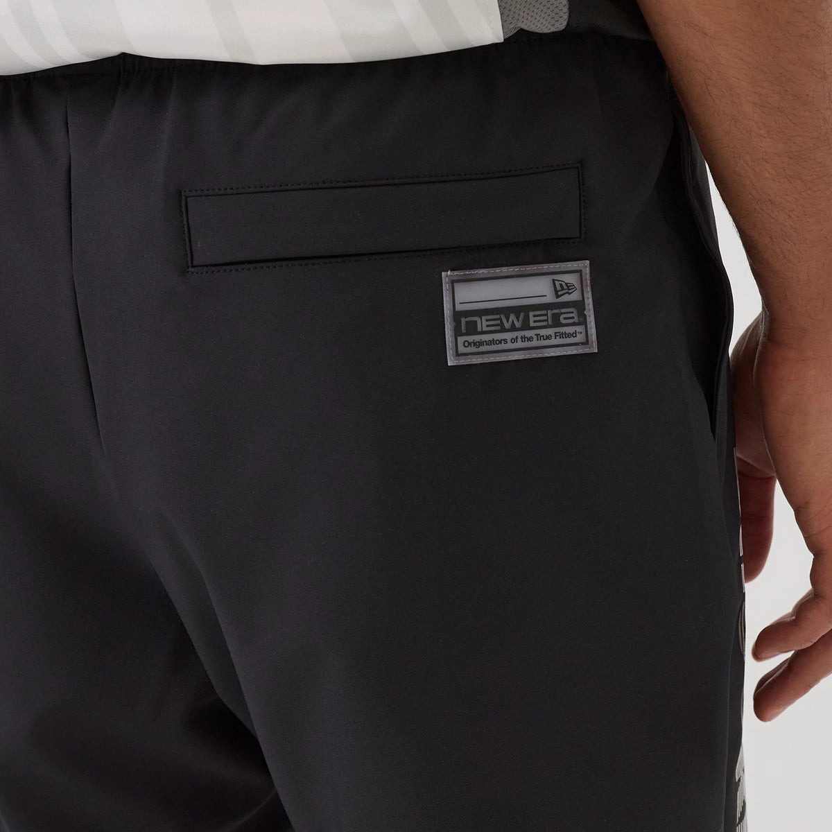 New Era Brooklyn Nets NBA Match Black Oversized Shorts, side view showing a rectangular grey patch with a debossed New Era logo near the waistband, a welt pocket slightly above, and a glimpse of vertical graphics on the side of the shorts.