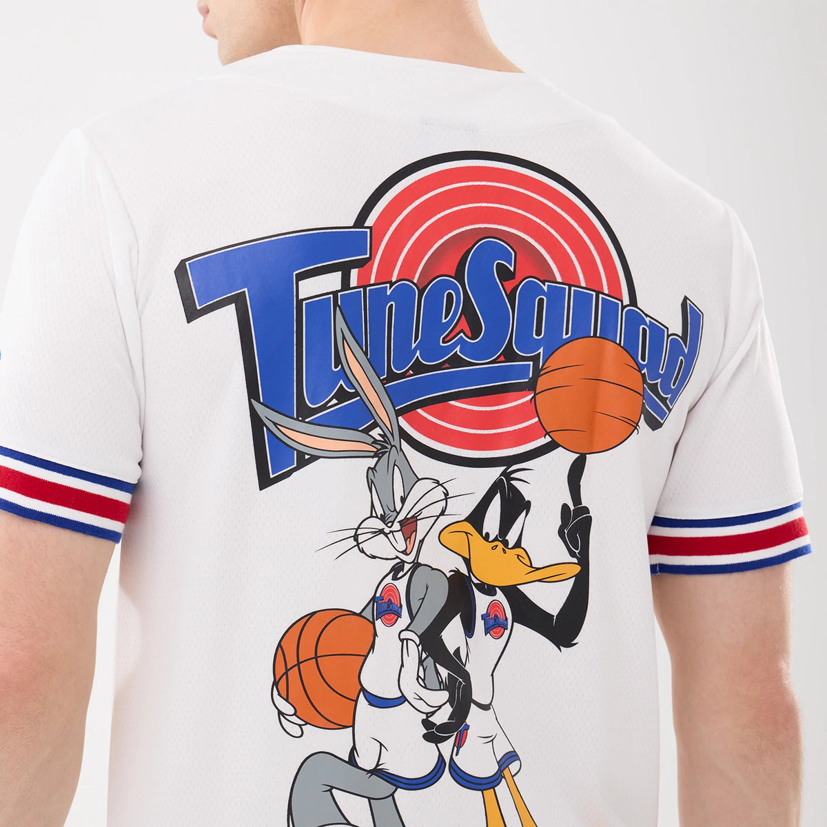 New Era Space Jam Warner Brothers Space Jam White Mesh Jersey, back view on a model showing Tune Squad lettering and graphics of Bugs Bunny and Daffy Duck holding basketballs on the white mesh fabric, with red and blue striped sleeves.