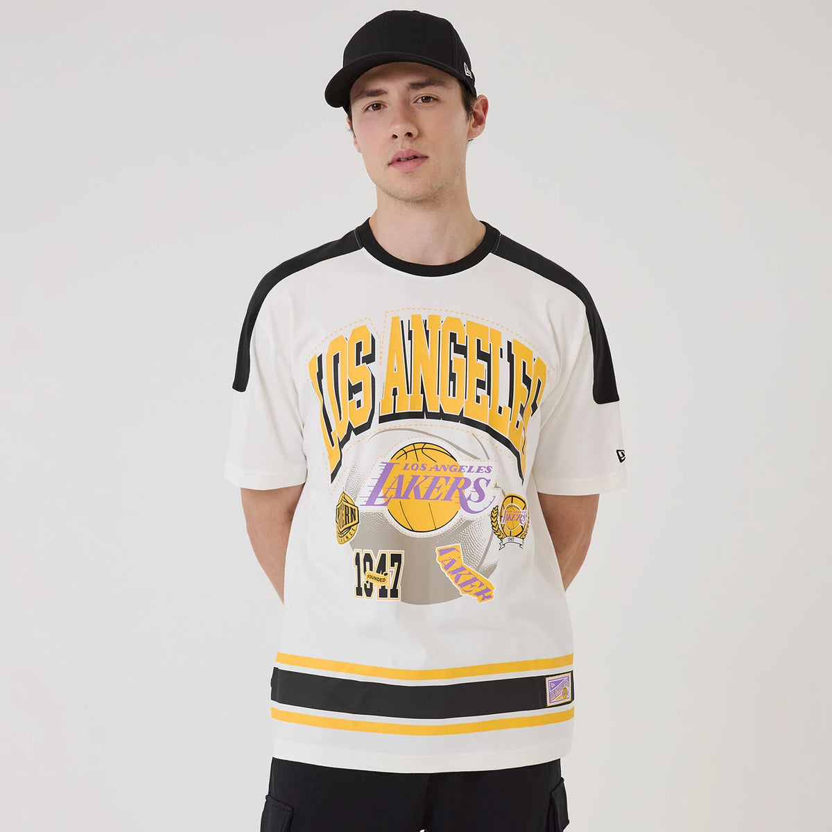 New Era LA Lakers NBA Sport Classic White T-Shirt, front view on-model showing large black and gold Los Angeles lettering across the chest with the Lakers logo below, yellow and black stripes along the hem, and embroidered New Era logo on the sleeve.
