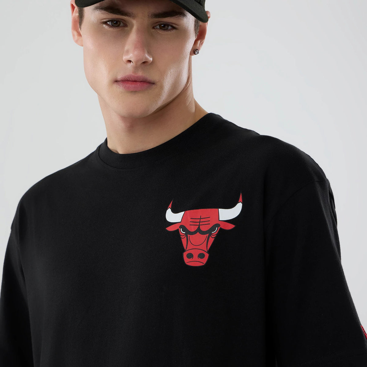 New Era Chicago Bulls NBA Logo Graphic Black T-Shirt, front view on-model displaying the red and white printed bull logo on the left chest of the black shirt, paired with a black cap.