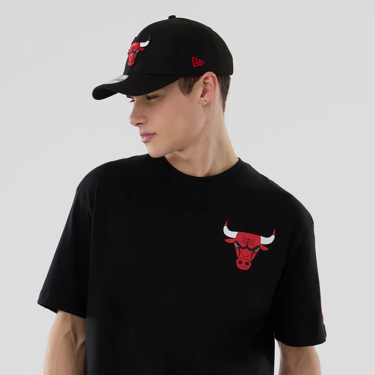New Era Chicago Bulls NBA Logo Graphic Black T-Shirt, front view on-model showing the red and white Bulls logo printed on the upper left chest, paired with a matching black cap featuring an embroidered logo on the front.