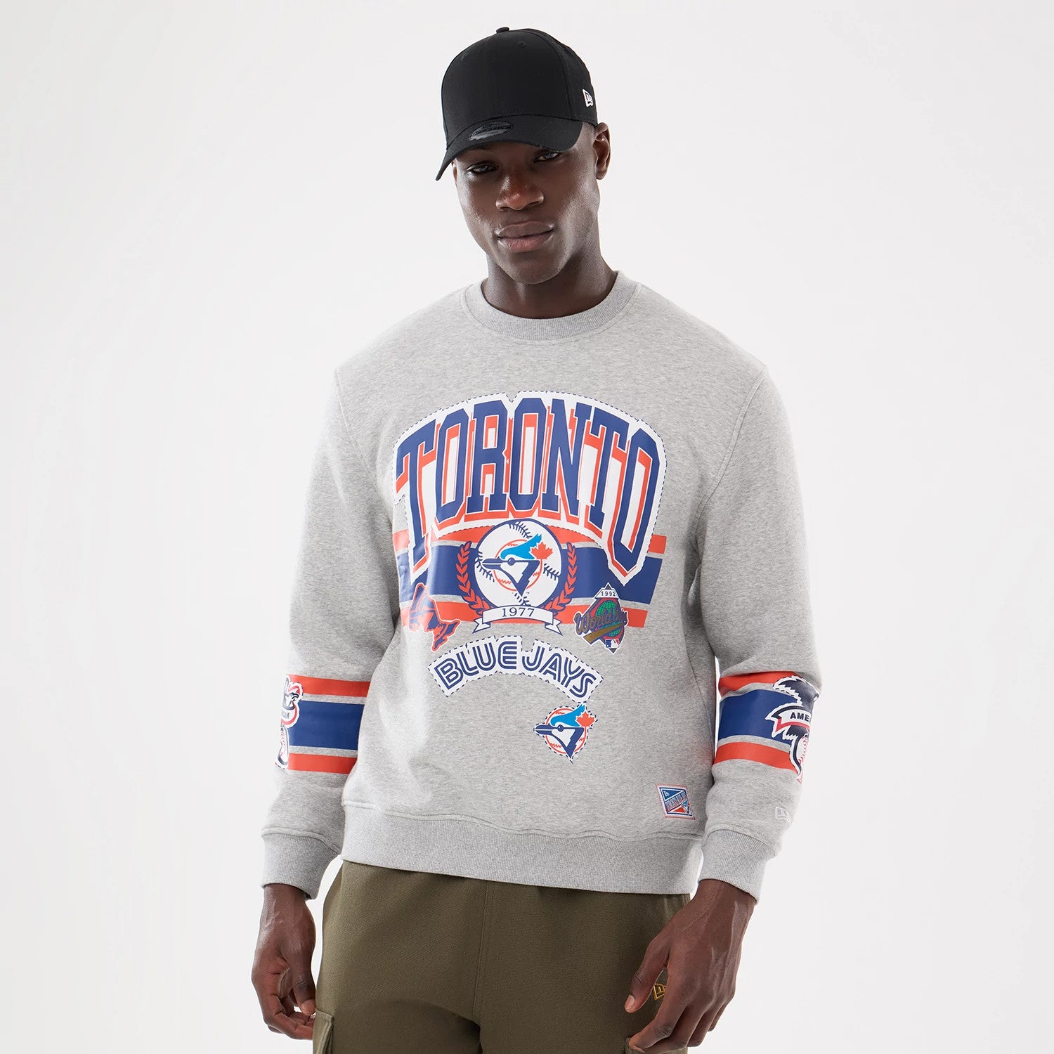 New Era Toronto Blue Jays MLB Sport Classic Grey Crew Neck Sweater, front view on a model with a black cap, featuring blue and orange Toronto lettering and Blue Jays logos on the chest and sleeves.