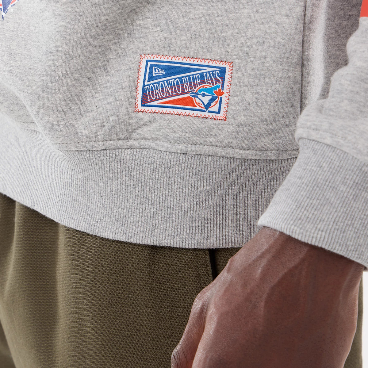 New Era Toronto Blue Jays MLB Sport Classic Grey Crew Neck Sweater, close-up showing the colourful patch with the New Era logo on the left hem, featuring the Blue Jays logo in red, blue and white embroidery.