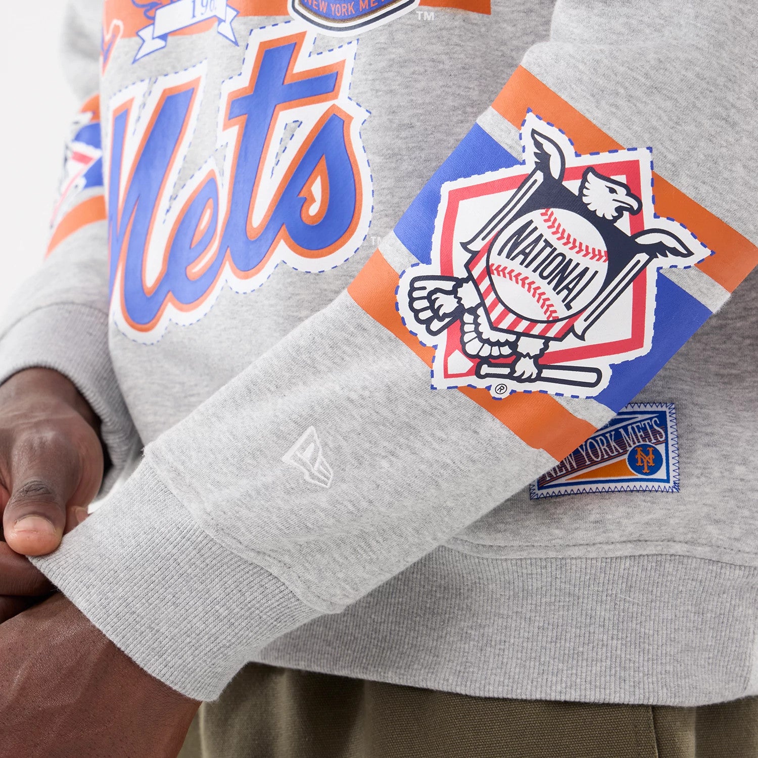New Era New York Mets MLB Sport Classic Grey Crew Neck Sweater, close-up showing the large Mets lettering on the front and a National League patch on the sleeve, with a white New Era flag logo embroidered on the cuff.