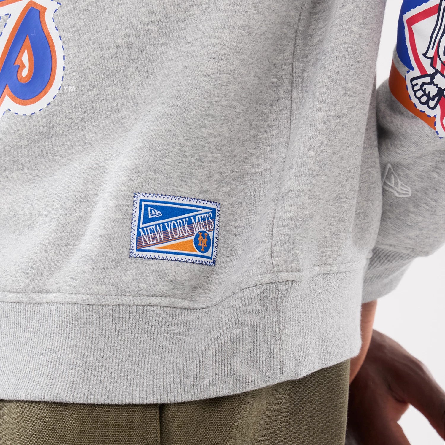 New Era New York Mets MLB Sport Classic Grey Crew Neck Sweater, side view showing an embroidered New York Mets patch near the ribbed hem and a stylised Mets graphic on the sleeve, with a small New Era flag logo.