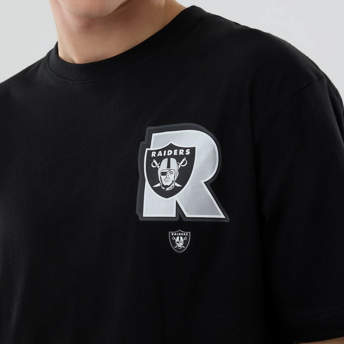 New Era Las Vegas Raiders NFL Graphic Black T-Shirt, front view showing a large grey R with the Raiders shield and helmeted figure on the chest, a smaller Raiders logo below, all on a plain black background.