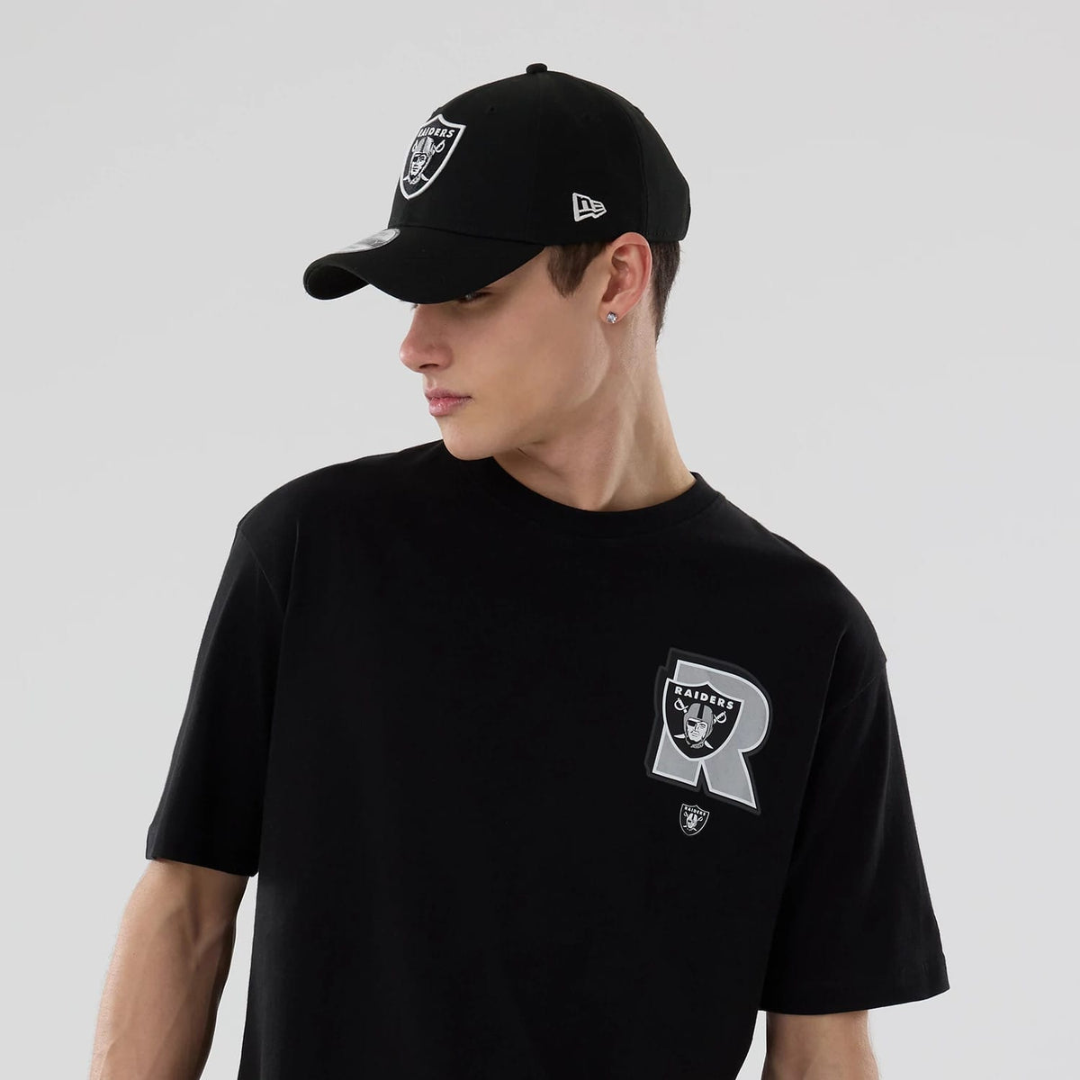 New Era Las Vegas Raiders NFL Graphic Black T-Shirt, front view on-model displaying an oversized grey and white Raiders logo beneath a large letter R on the chest, paired with a black cap featuring an embroidered Raiders logo on the front.