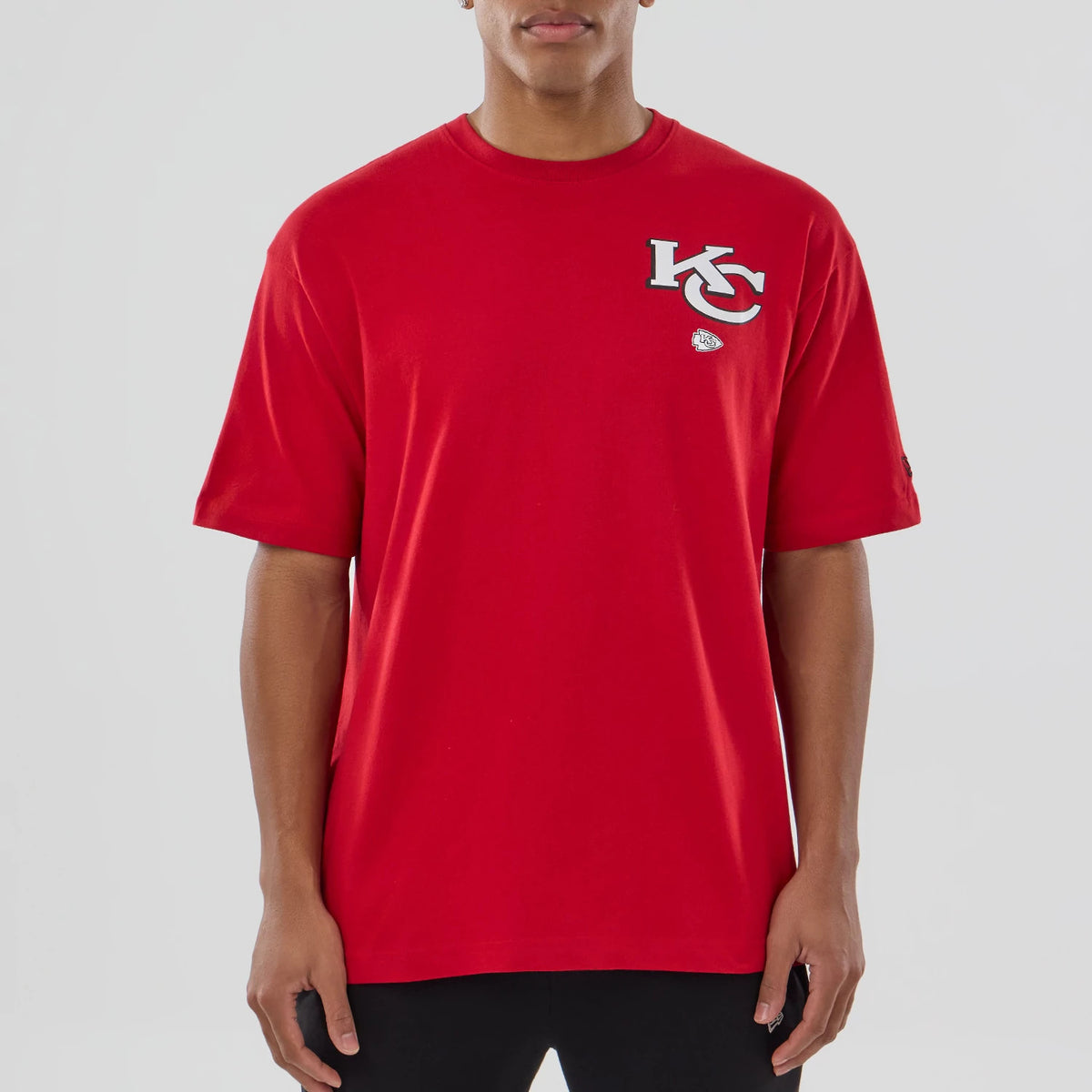 New Era Kansas City Chiefs NFL Graphic Red T-Shirt, front view on-model displaying a large white embroidered KC logo above a smaller embroidered football emblem on the chest, with short sleeves and a plain red design.