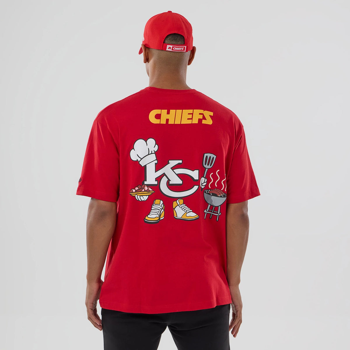 New Era Kansas City Chiefs NFL Graphic Red T-Shirt, back view on a model with a red cap, featuring large yellow Chiefs lettering at the top and playful graphic elements including a KC logo, chef hat, and barbecue design across the back.