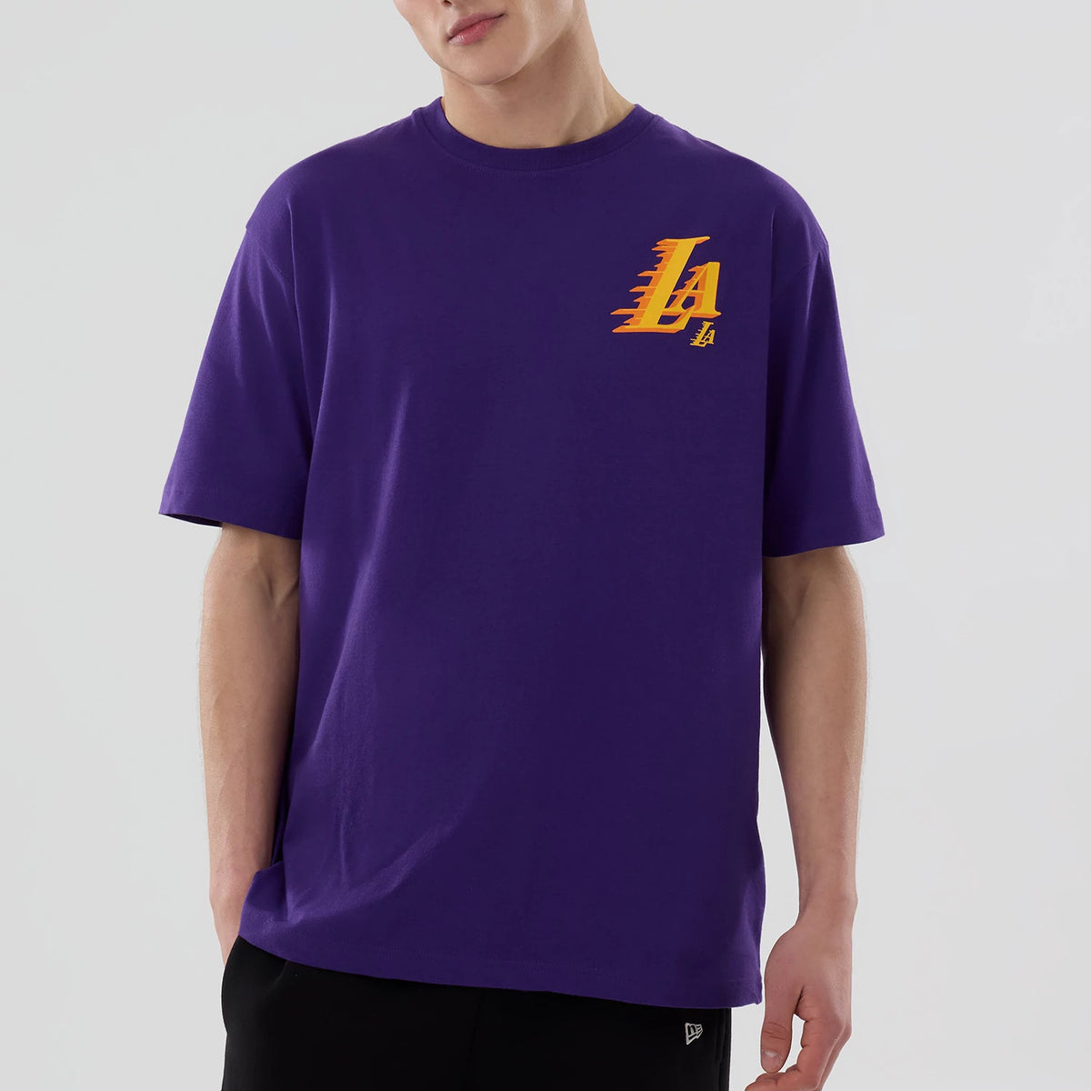 New Era LA Lakers NBA Graphic Purple T-Shirt, front view on-model showing a yellow and orange LA Lakers logo printed on the left chest of the purple tee, styled with plain black trousers.