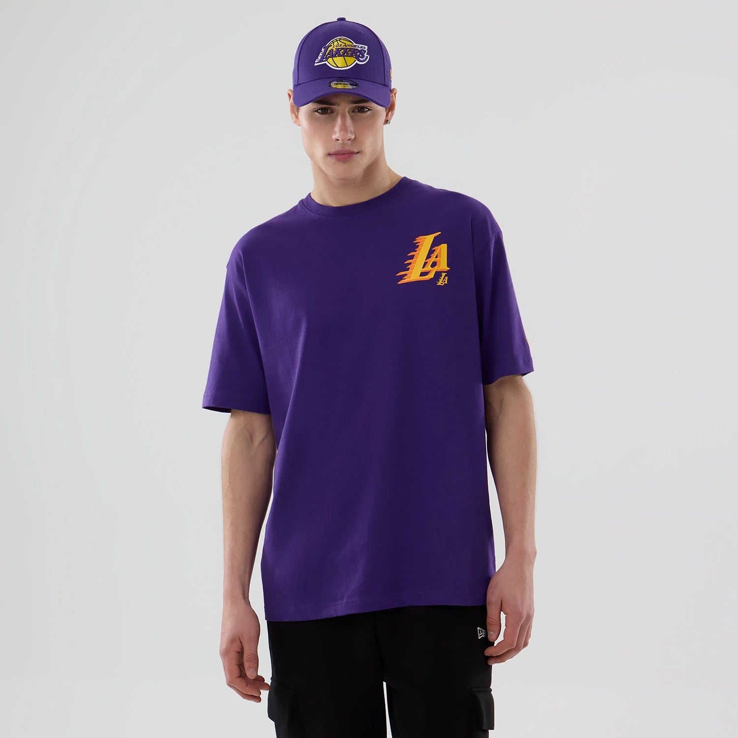New Era LA Lakers NBA Graphic Purple T-Shirt, front view on-model showing the yellow and orange LA logo on the left chest, styled with a matching cap featuring an embroidered Lakers emblem on the front.