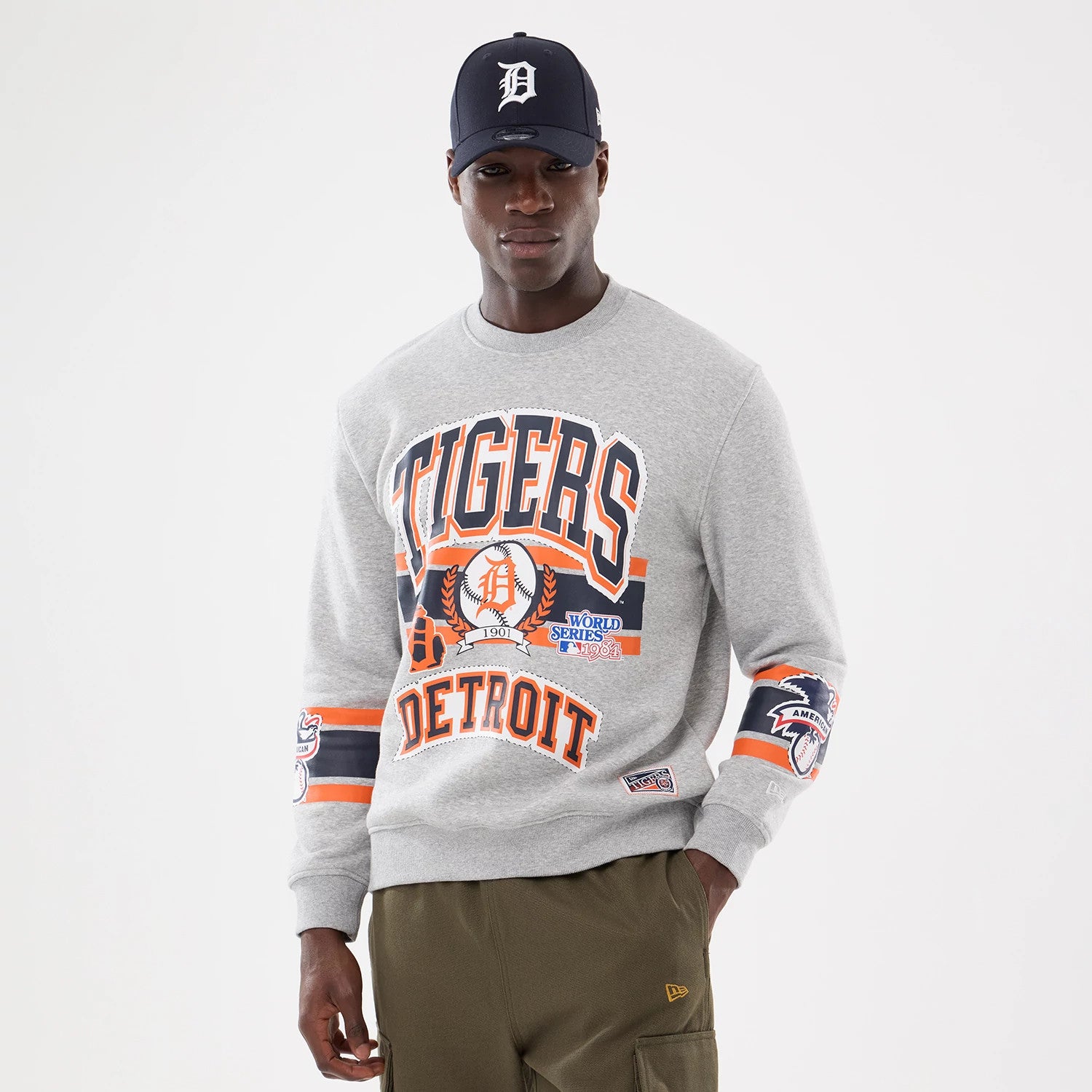 New Era Detroit Tigers MLB Sport Classic Grey Crew Neck Sweater, front view on a model wearing a navy cap, featuring orange and navy lettering with team logos on the chest and sleeve, complemented by orange-striped cuffs.