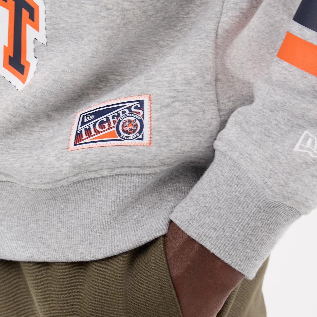 New Era Detroit Tigers MLB Sport Classic Grey Crew Neck Sweater, close-up showing the detailed embroidered Tigers patch on the lower hem, with ribbed cuffs and a New Era flag logo on the sleeve.