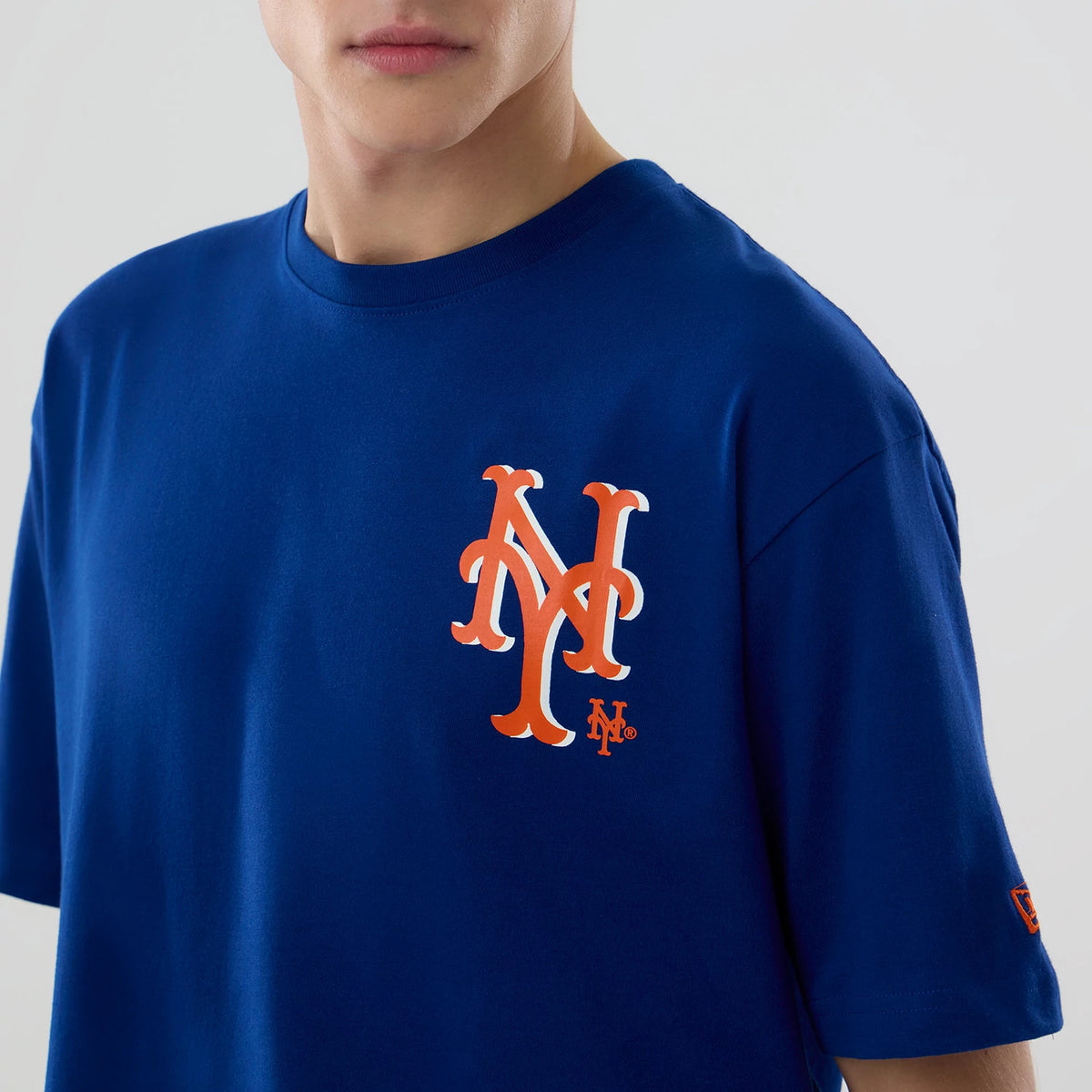 New Era New York Mets MLB Graphic Blue T-Shirt, front view on-model showing a large orange printed NY Mets logo on the left chest and a small embroidered New Era logo on the left sleeve.