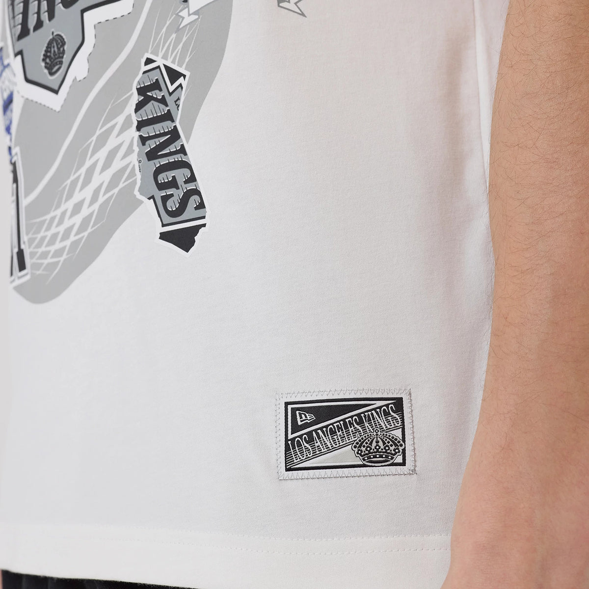 New Era LA Kings NHL Sport Classic White T-Shirt, side view showing the lower edge with a black embroidered Los Angeles Kings patch near the hem and various graphic elements in black and grey on the white fabric.