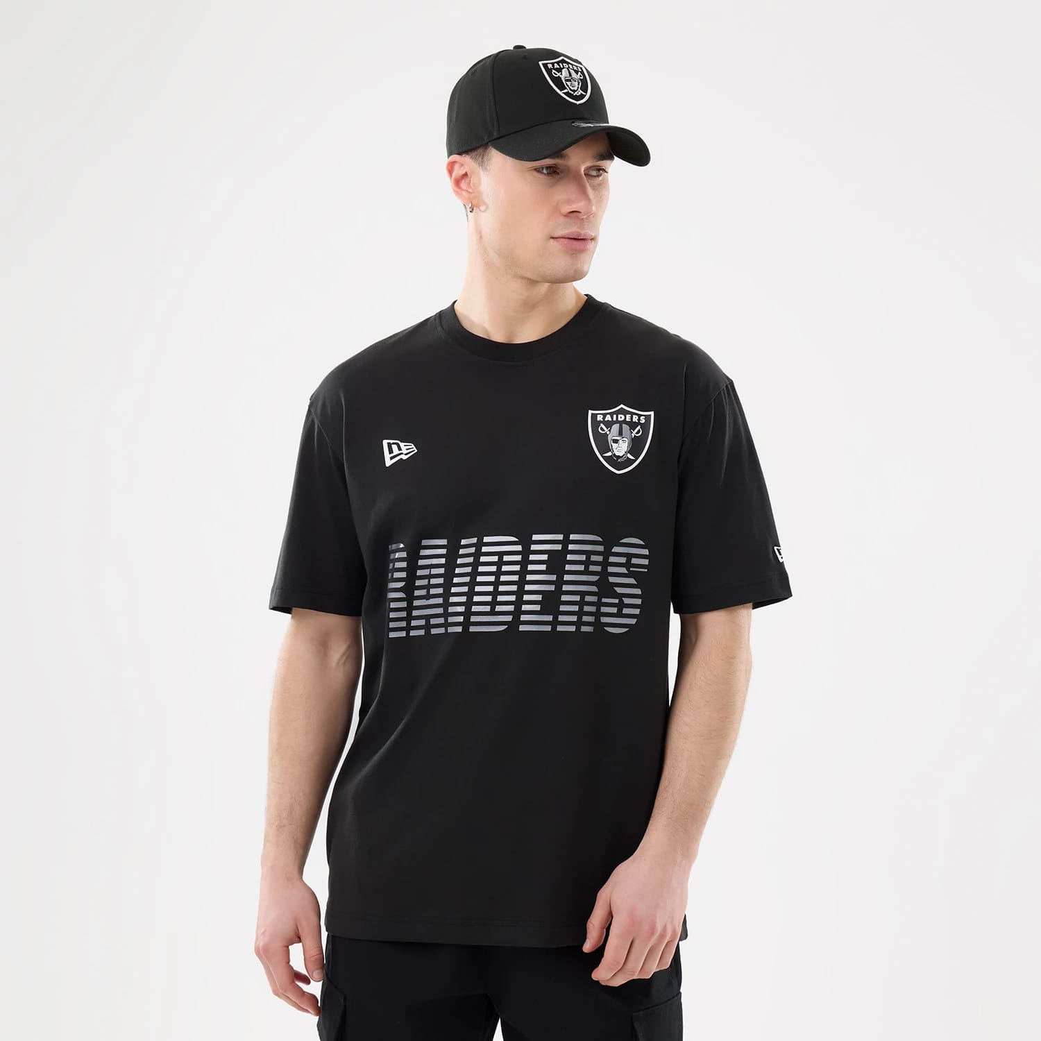 New Era Las Vegas Raiders NFL Match Black T-Shirt, front view on a model wearing a matching cap, showing a raised Raiders shield logo on the chest, a printed Raiders wordmark below, and an embroidered New Era logo on the right.