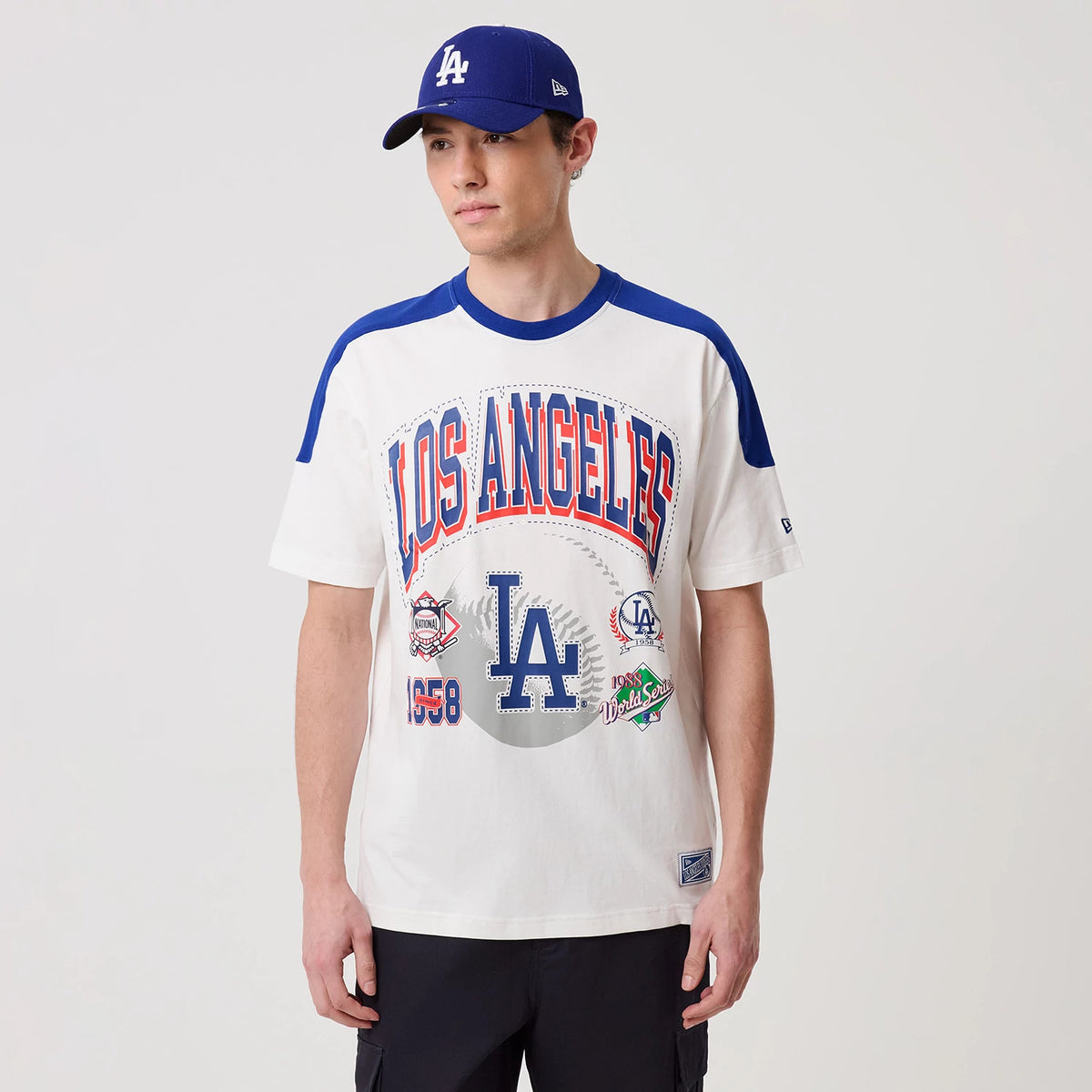 New Era LA Dodgers MLB Sport Classic White T-Shirt, front view on a model showcasing the Los Angeles lettering in red and blue across the chest, a large blue LA logo, and various baseball graphics with a blue collar and shoulder detail.