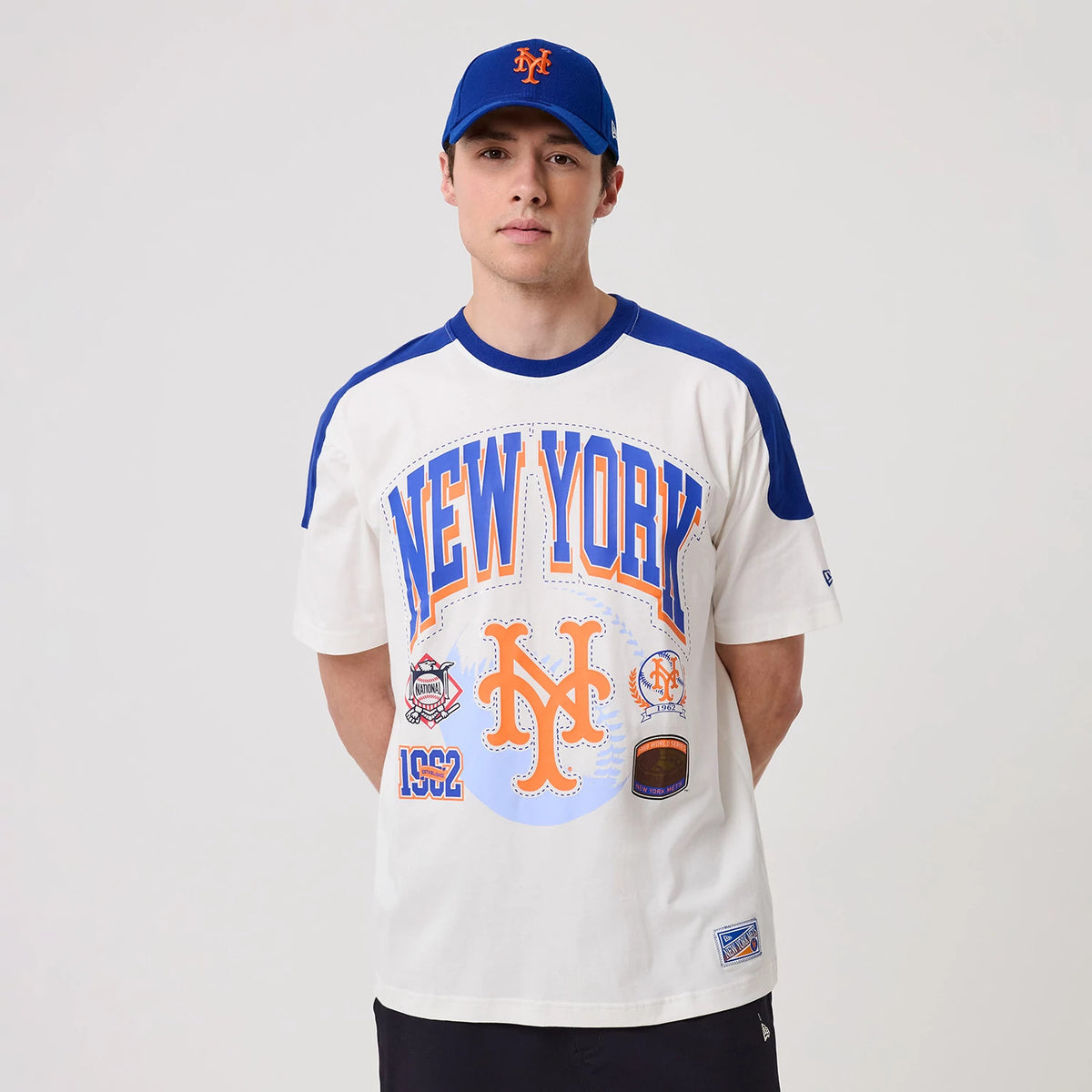 New Era New York Mets MLB Sport Classic White T-Shirt, front view on a model paired with a matching blue cap, featuring large blue and orange New York lettering, a printed Mets logo, and decorative patches across the chest.