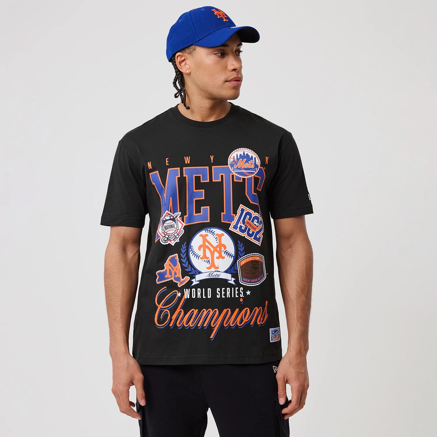 New Era New York Mets MLB Sport Classic Black T-Shirt, front view featuring Mets and Champions graphics in blue and orange across the chest, worn by a model with a matching blue cap.