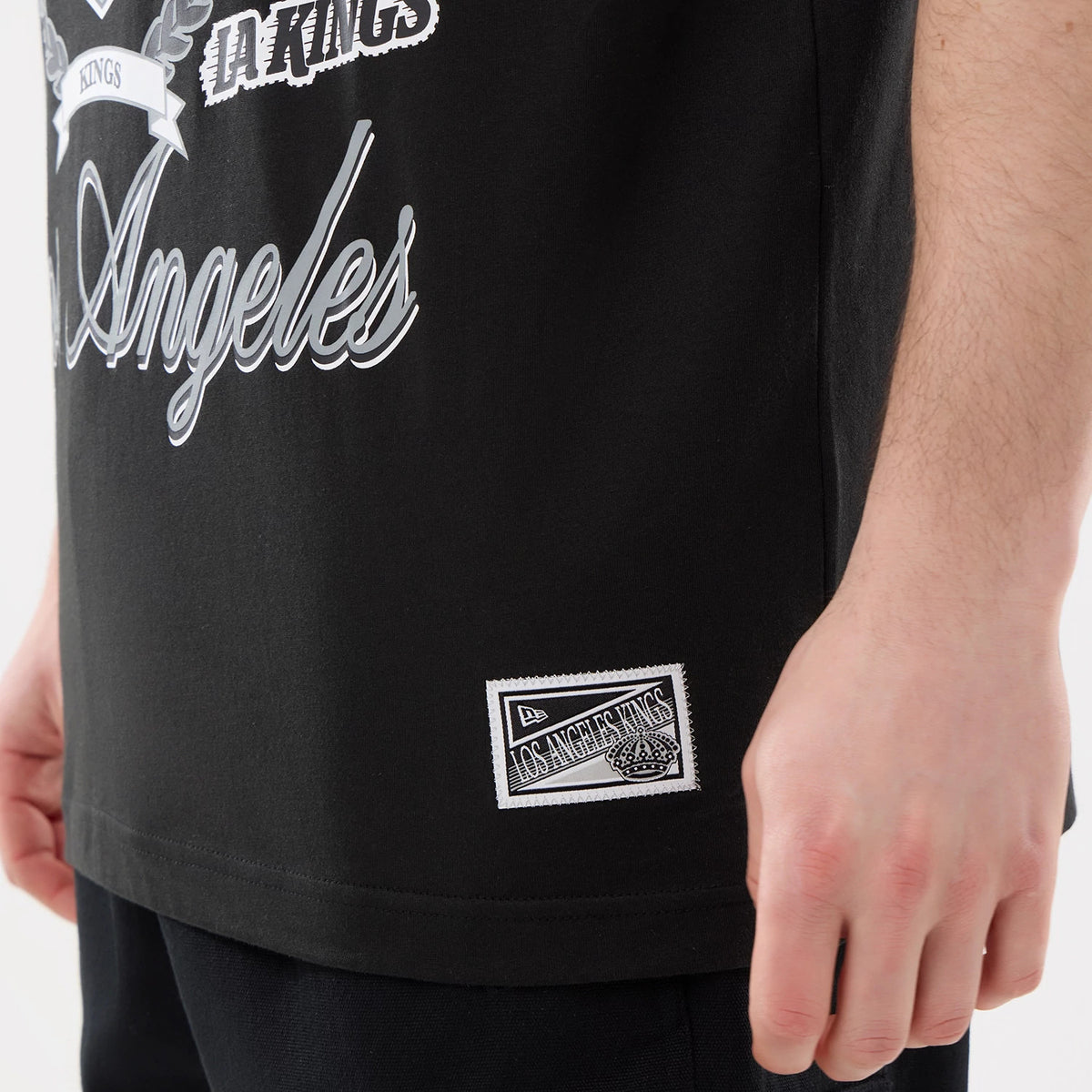 New Era LA Kings NHL Sport Classic Black T-Shirt, side view showing detailed white graphic lettering on the black fabric with an embroidered patch at the lower hem, styled casually with dark trousers on-model.