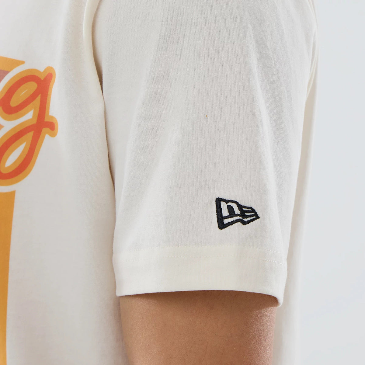 New Era Cactus League Logo MLB Spring Training 2026 Cream T-Shirt, side view showing the short sleeve with a black embroidered New Era logo near the cuff and the large orange graphics continuing across the chest.