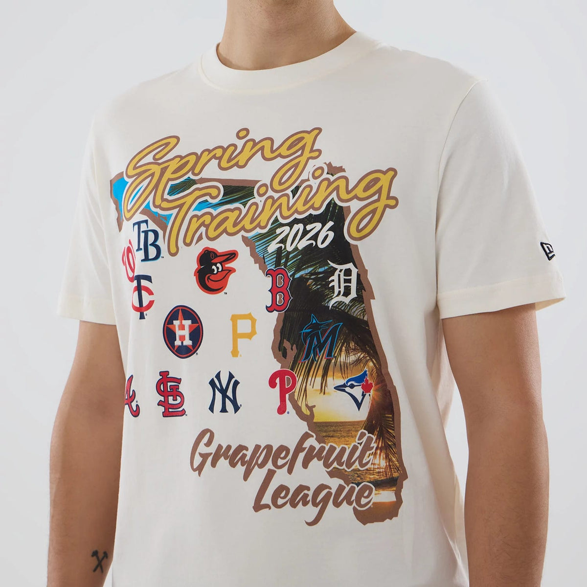 New Era Grapefruit League Logo MLB Spring Training 2026 Cream T-Shirt, front view showing graphics of the words Spring Training and Grapefruit League with various team logos, and an embroidered New Era logo on the sleeve.