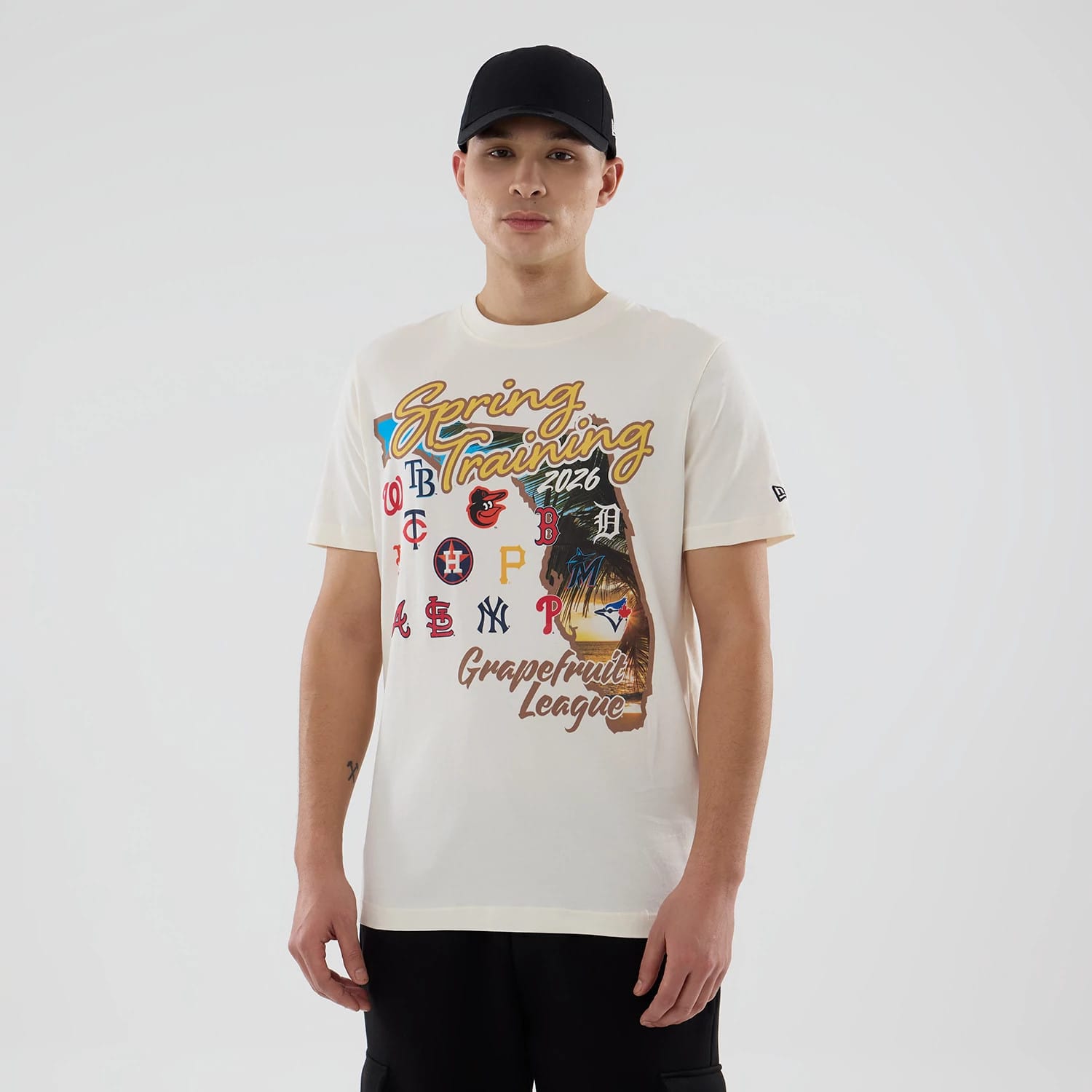 New Era Grapefruit League Logo MLB Spring Training 2026 Cream T-Shirt, front view on a model featuring multiple MLB team logos, large Spring Training lettering and a graphic of a player, with a black New Era cap.