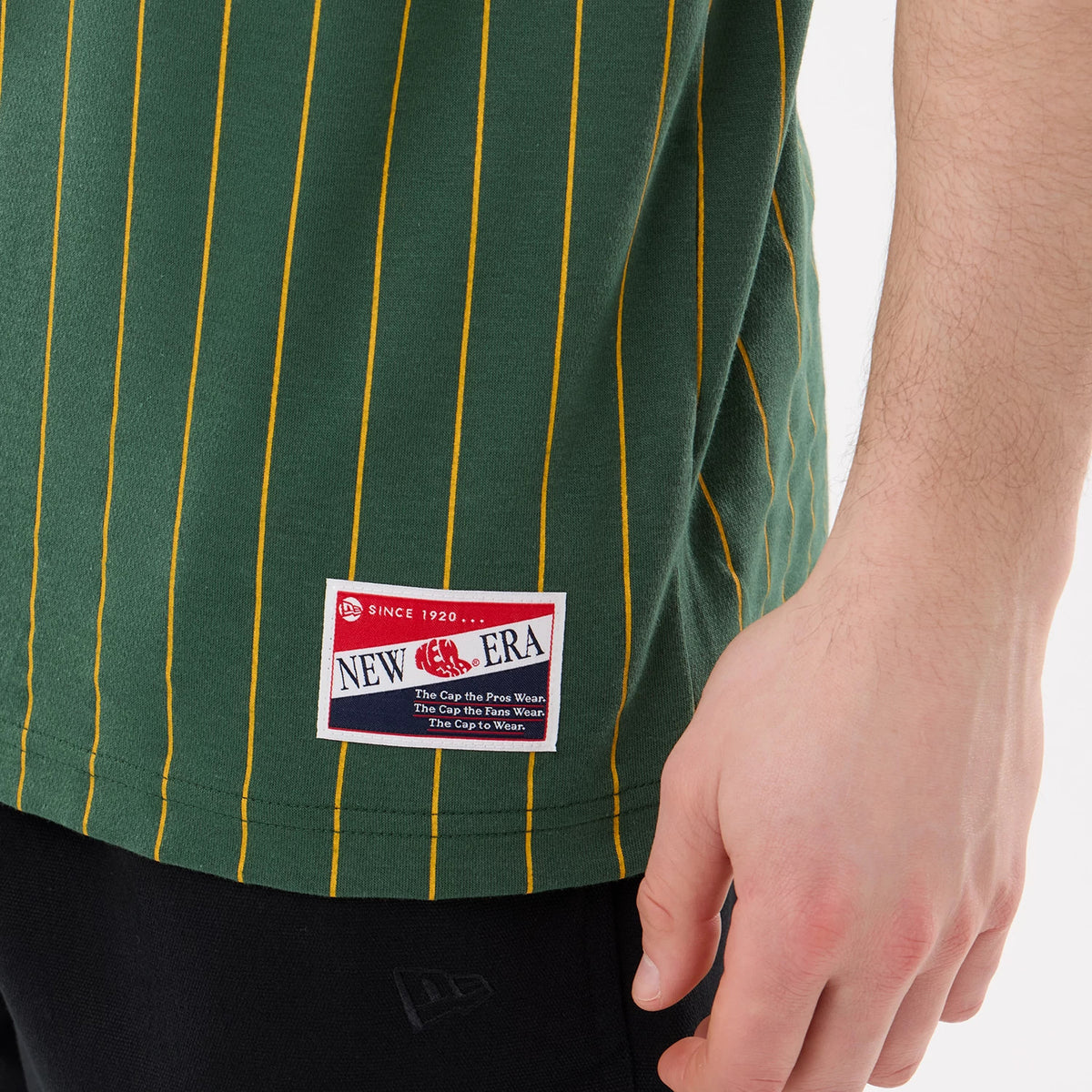 New Era Athletics MLB Pinstripe Dark Green T-Shirt, side view showing narrow yellow pinstripes on dark green fabric with a rectangular New Era embroidered patch near the bottom hem, worn with black trousers.