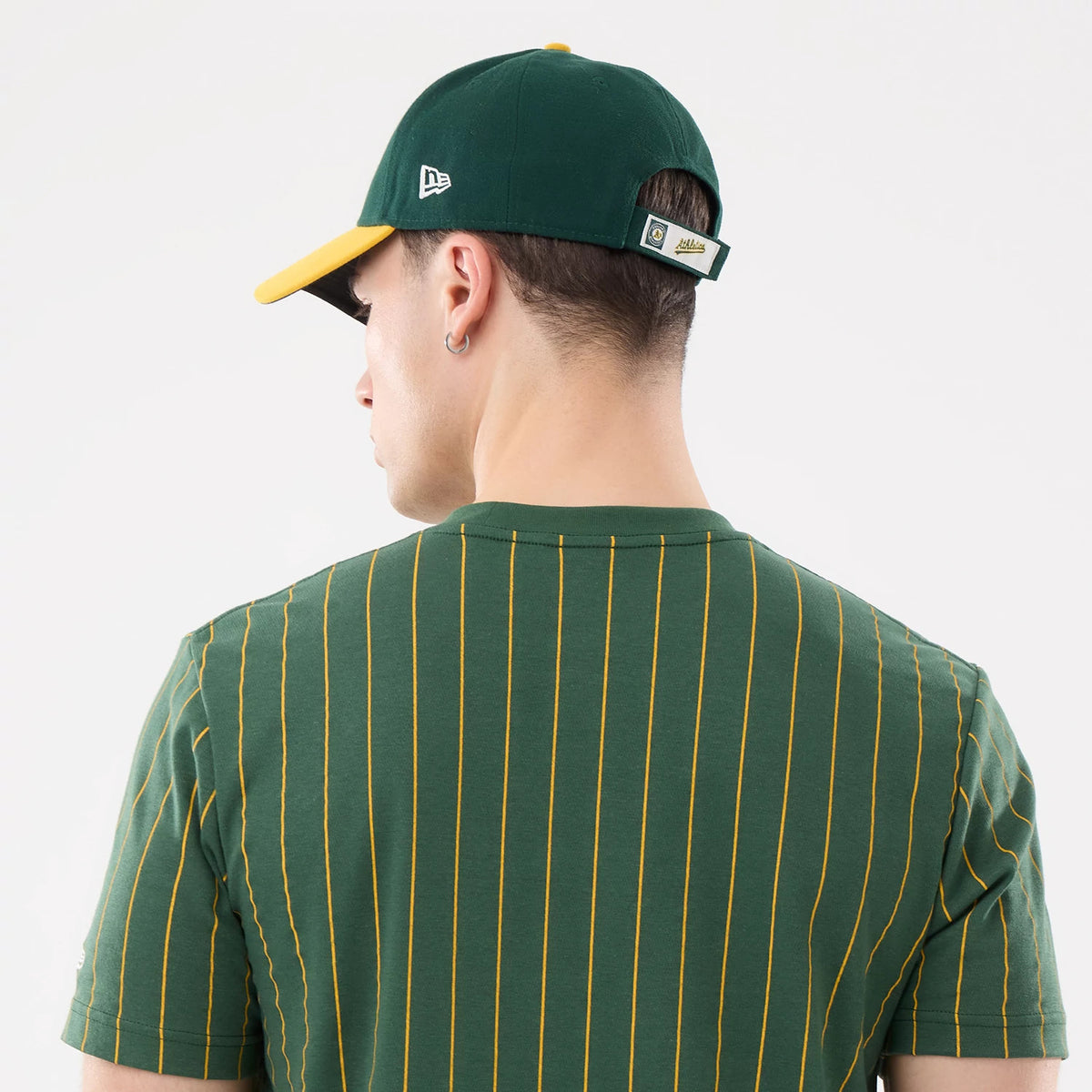 New Era Athletics MLB Pinstripe Dark Green T-Shirt, back view on a model with yellow pinstripes running vertically, paired with a dark green cap featuring an embroidered New Era logo on the side and a yellow underside on the visor.